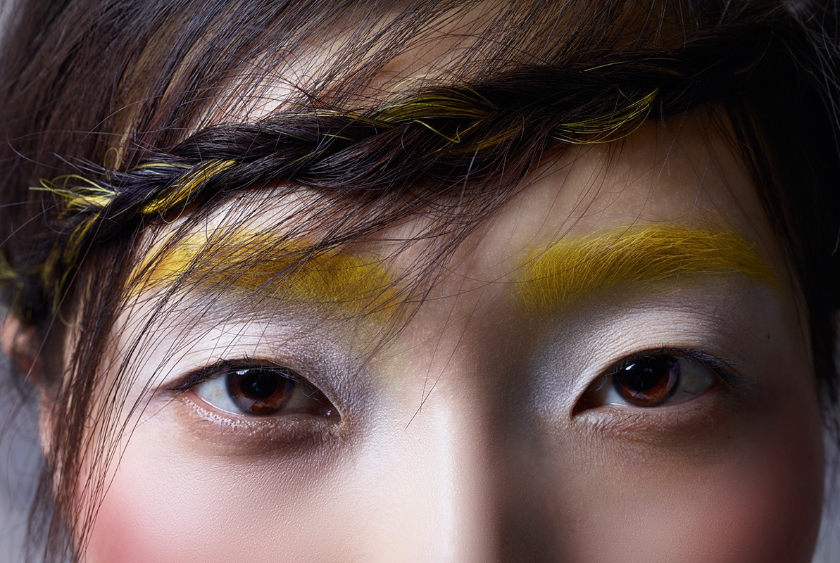 Close-up of a woman with brown eyes, yellow eyebrows and braided hair, wearing white eye shadow and pink blush against a grey background.