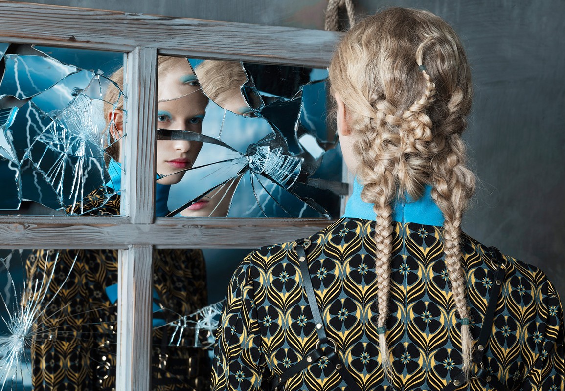Fashion shot with blonde woman facing away, showcasing braided hair, patterned jacket, and broken mirror reflecting a face with blue eyeshadow.