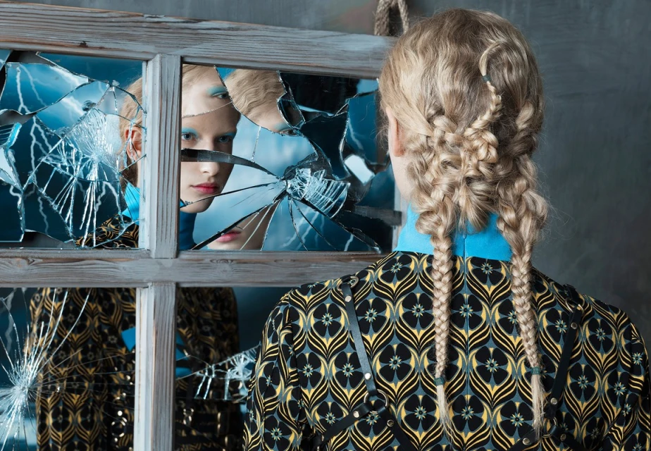Fashion shot with blonde woman facing away, showcasing braided hair, patterned jacket, and broken mirror reflecting a face with blue eyeshadow.
