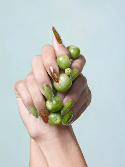 Close-up of a woman's hands decorated with creative nail art featuring tomatillos against a pale blue background in studio lighting.