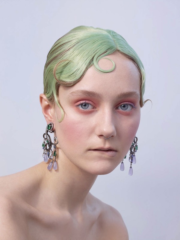 Close-up of a fair-skinned young woman with light green, wavy hair, wearing large ornate earrings with purple and green gems against a light background.