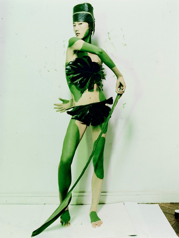 Asian female model with green body paint and leaf-based headdress and outfit stands on white backdrop in a studio setting.