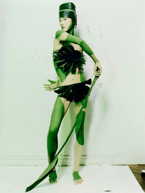 Asian female model with green body paint and leaf-based headdress and outfit stands on white backdrop in a studio setting.