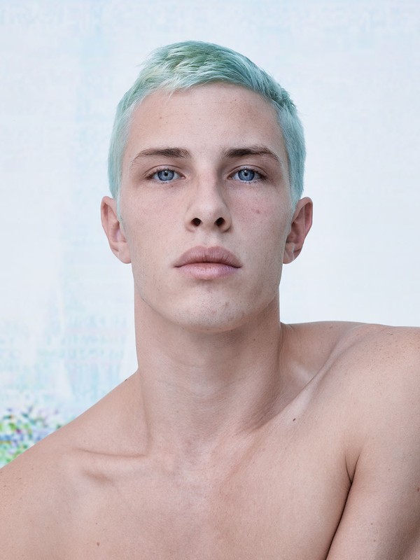Portrait of a shirtless adult male with blue eyes and dyed light-blue hair, posing indoors against a blurred, light-colored background.