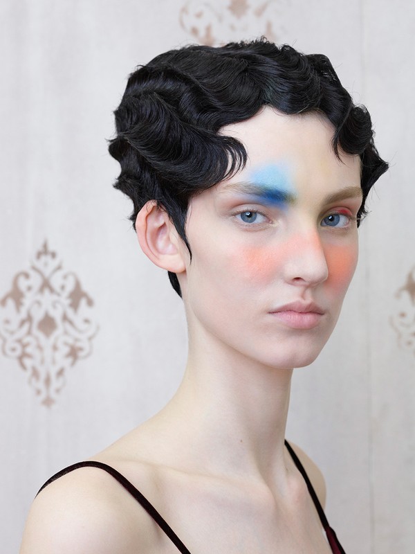 Close-up portrait of a person with short, dark, wavy hair and fair skin, featuring colorful makeup on the face against a patterned backdrop.