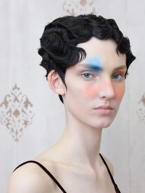 Close-up portrait of a person with short, dark, wavy hair and fair skin, featuring colorful makeup on the face against a patterned backdrop.
