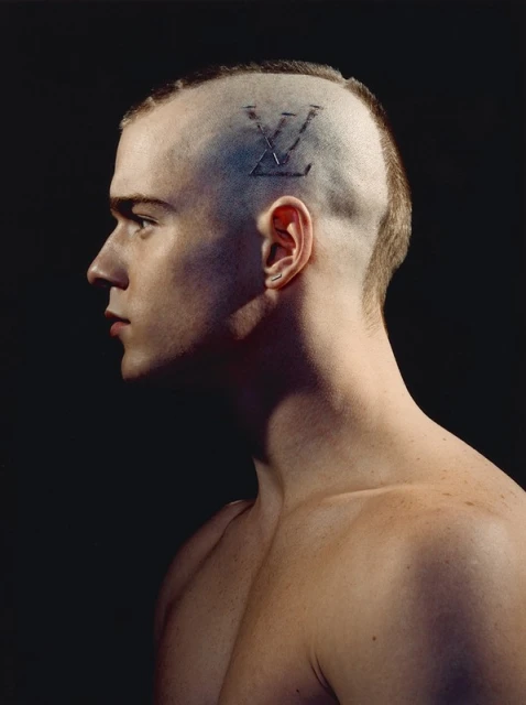 Profile of a young adult male with shaved head and a logo design, set against a dark background.  Upper body is visible.