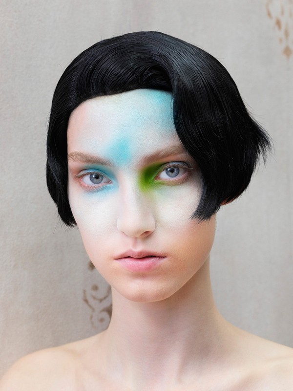 Close-up portrait of a fair-skinned person with black hair and striking makeup featuring blue and green hues, against a neutral backdrop.