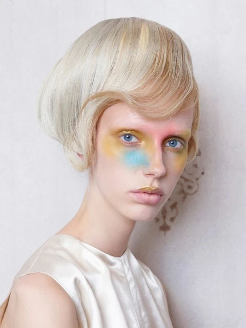 Striking beauty shot of a young woman with a blonde bowl cut, fair skin, and creative colorful makeup in a studio setting.