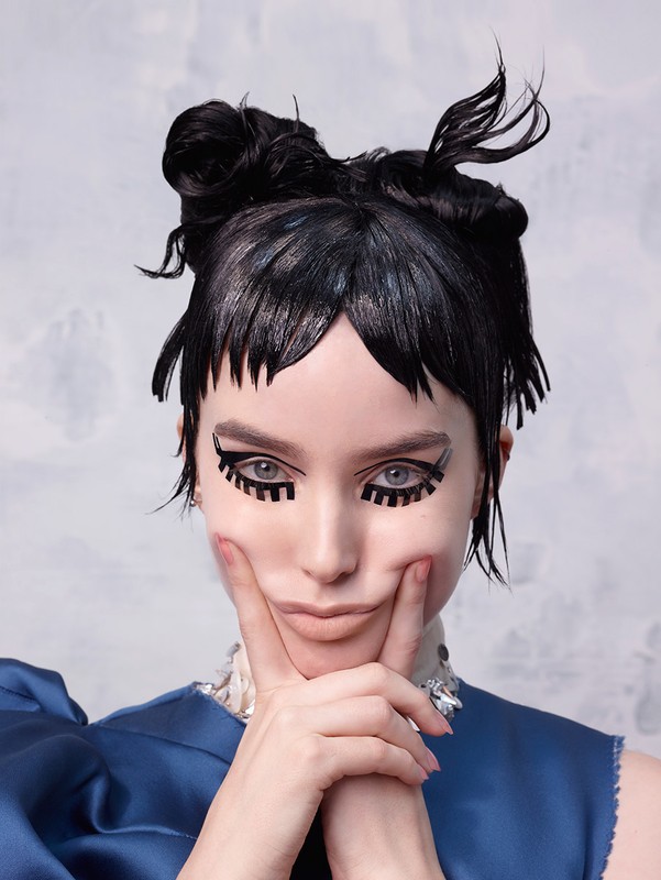 Adult female poses with creative eye makeup, dark double-bun hairstyle, wearing a blue dress in front of a patterned light blue background.