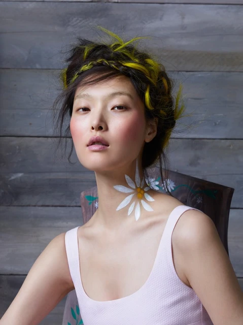 Portrait of an Asian woman with yellow highlights, daisy body art, and a pink top, seated against a weathered wooden backdrop. Beauty shoot.