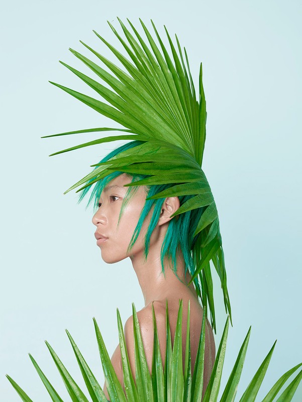 Profile of a person with green hair and makeup, adorned with palm leaves against a light blue backdrop in a studio shot.