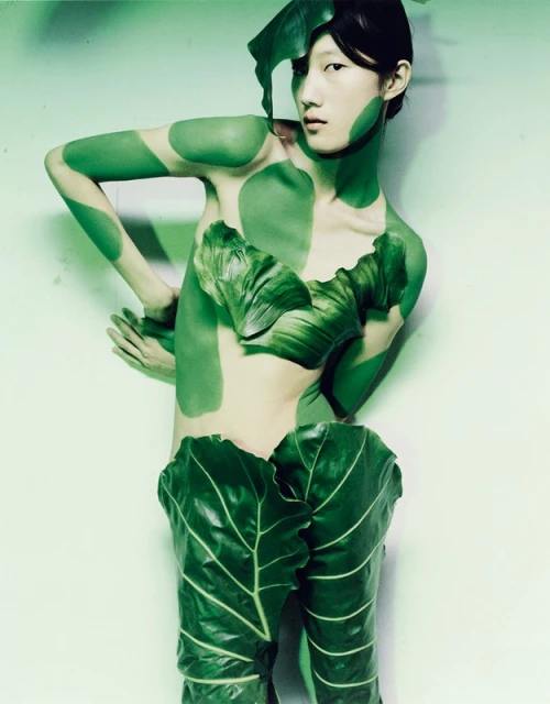 Young woman in conceptual fashion shot, posed in partial body paint with large green leaves as clothing against a plain background.