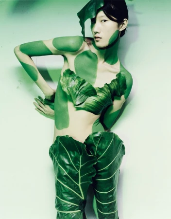 Young woman in conceptual fashion shot, posed in partial body paint with large green leaves as clothing against a plain background.