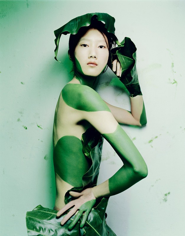 Asian woman with green body paint and leaf clothing posing against a light green wall in a portrait-style artistic photograph.