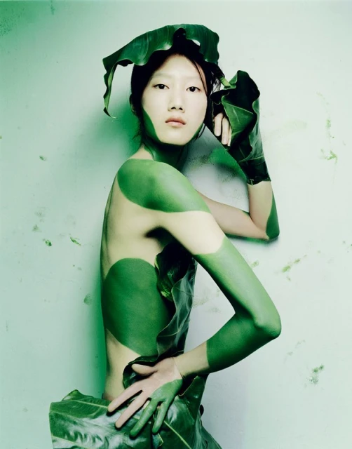 Asian woman with green body paint and leaf clothing posing against a light green wall in a portrait-style artistic photograph.