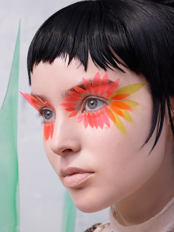 Close-up shot of a woman with creative eye makeup, featuring colorful petal-like designs, black hair, and fair skin, in a beauty style.
