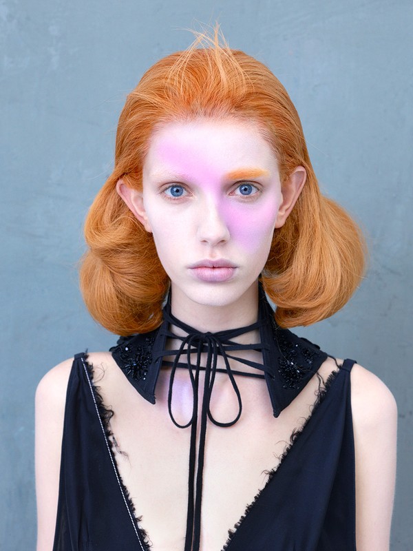 Close-up portrait of a young woman with red hair and pale skin wearing a black choker-style collar and a black dress against a grey background.