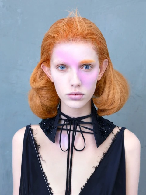 Close-up portrait of a young woman with red hair and pale skin wearing a black choker-style collar and a black dress against a grey background.