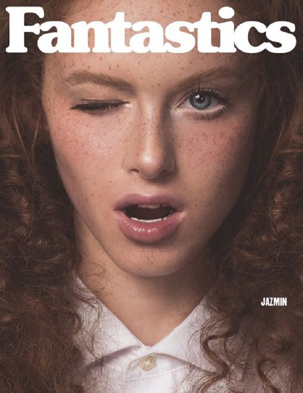 Close-up portrait of a young woman with freckles winking with one eye, styled in a white collared shirt, against a dark background.