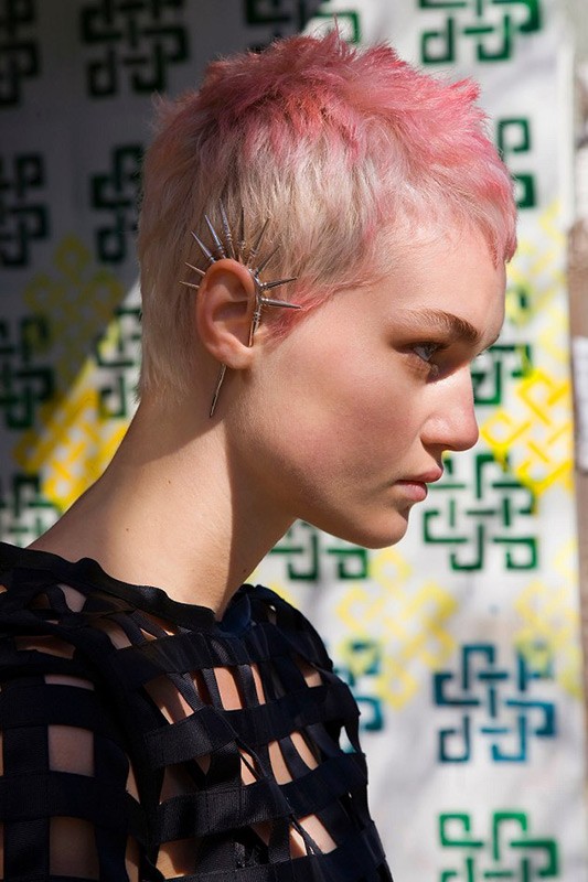 Close-up of a young adult woman with a light complexion, pink hair, and ear jewelry, wearing a black patterned top against a geometric backdrop.