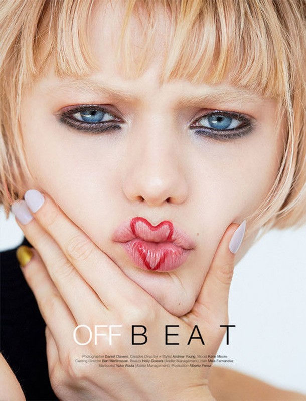 Close-up of a young woman with blonde hair, blue eyes, fair skin, and a red heart painted on her lips, holding her hand to her face.