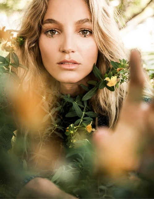 Portrait of a blonde woman with yellow flowers in her hair and around her face, set against a natural outdoor background.