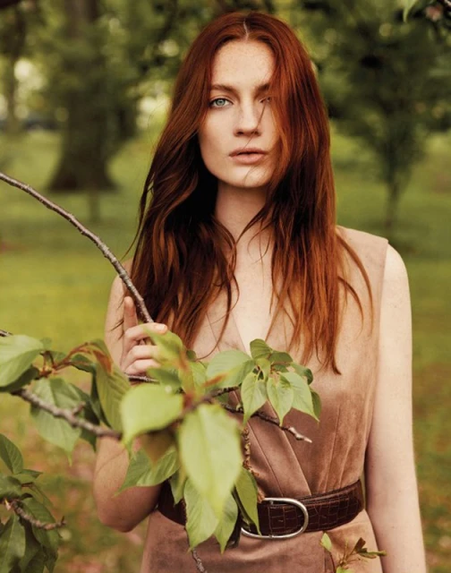 Red-haired woman in earthy-toned dress poses in nature with a leafy branch in foreground, soft daylight, creating a natural fashion portrait.