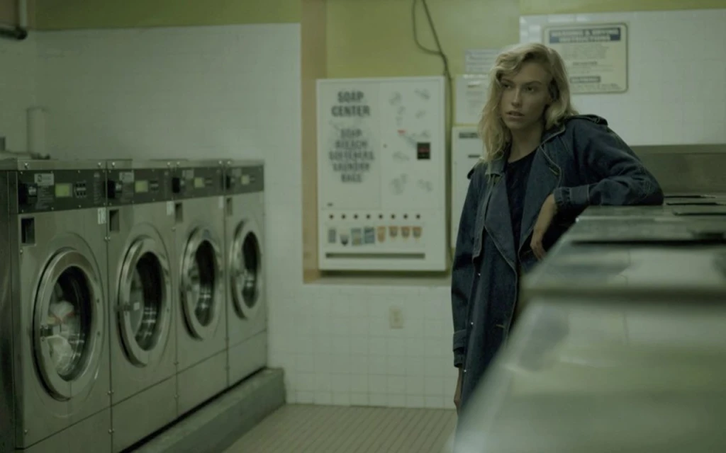 Blonde woman in denim coat in a dimly lit laundromat with chrome washing machines and a soap vending machine in the background.