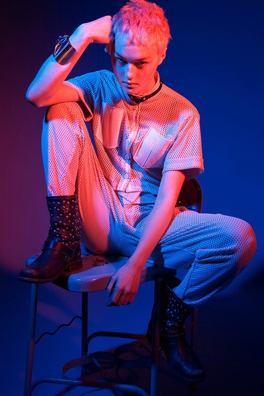 An adult with pink hair is sitting on a stool in a studio wearing a mesh jumpsuit and studded boots with red and blue lighting.