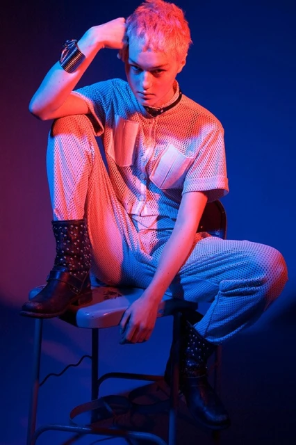 An adult with pink hair is sitting on a stool in a studio wearing a mesh jumpsuit and studded boots with red and blue lighting.