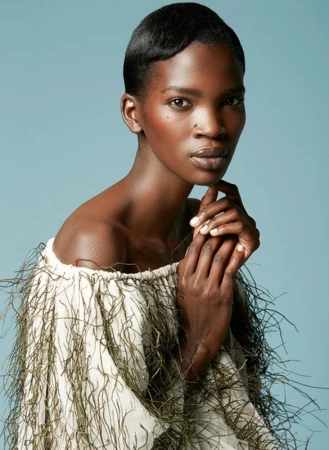 Close-up studio shot of a woman with dark skin and short hair, wearing an off-the-shoulder top with fringe, against a pale blue background.