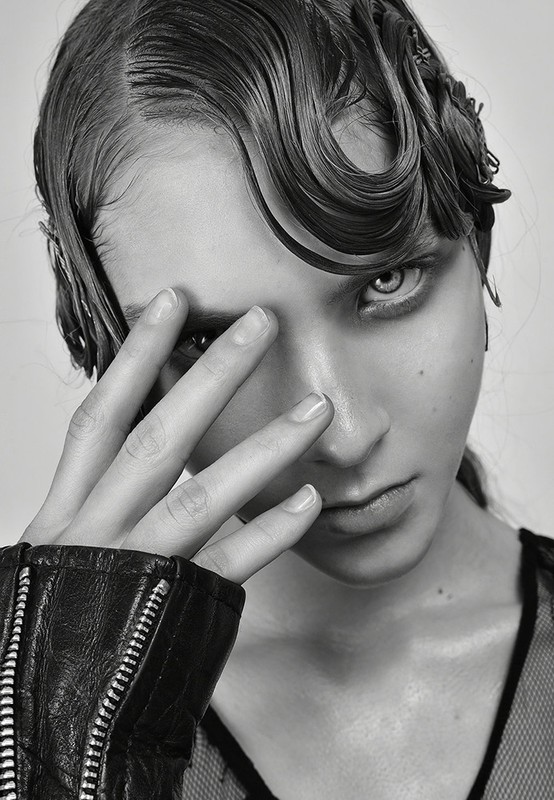 Close-up portrait of a model with wet-styled hair and hand raised across face, wearing a black leather jacket.