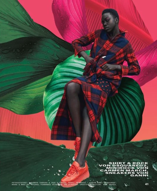 A slender, dark-skinned woman poses elegantly in a red and blue plaid outfit and orange sneakers against a backdrop of pink and green leaves.