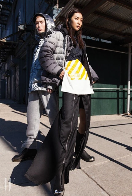 Fashion shoot featuring an Asian man and woman modeling streetwear puffer jackets on a city street with an industrial background.
