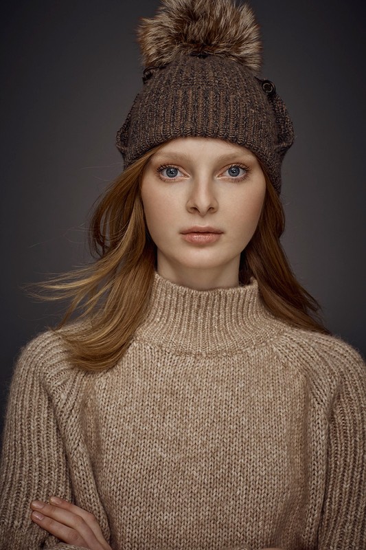 Portrait of a young woman with red hair wearing a knitted hat with fur pompom and a beige turtleneck sweater against a gray background.