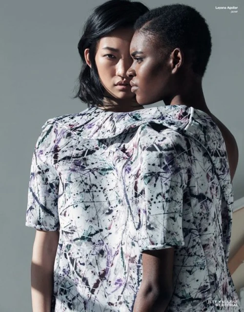 Two women with dark hair are closely posed in front of a light gray studio backdrop. The woman in the front wears a patterned top.