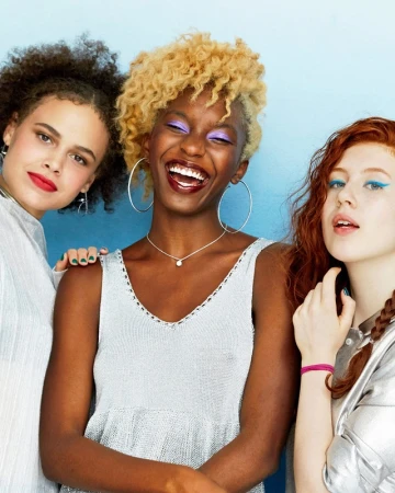 Three women with vibrant makeup posing indoors. The central woman smiles broadly, showcasing purple eyeshadow and a silver top, in portrait.