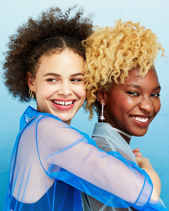 Two smiling young women of different ethnicities pose together indoors against a blue background wearing sheer shirts and earrings.