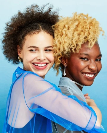 Two smiling young women of different ethnicities pose together indoors against a blue background wearing sheer shirts and earrings.