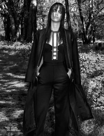 Monochrome fashion portrait of a model with dark wet-look hair wearing a black jacket and trousers in a forest.