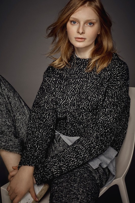 A fair-skinned young woman in a textured knit sweater sits in a photo studio. She is fair skinned with reddish blonde hair.