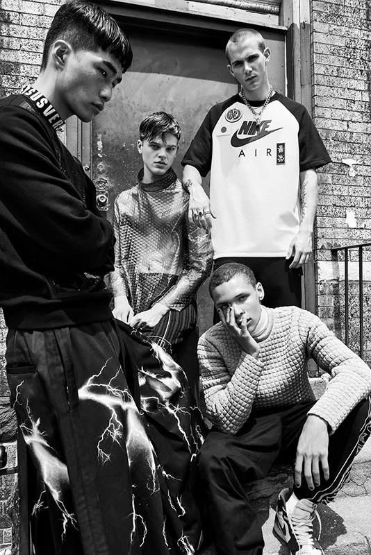 A group of four young adult men pose outdoors in casual streetwear fashion against a brick building in black and white.