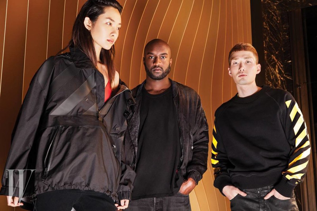 Three fashion models in black Off-White branded jackets and sweatshirt pose against a golden, curved striped backdrop. 