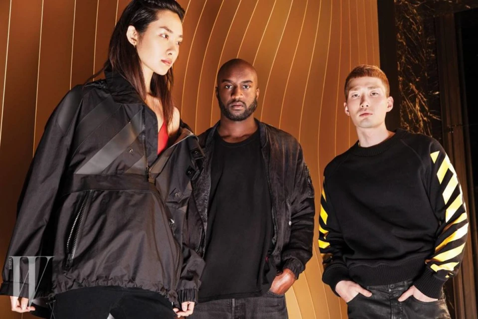 Three fashion models in black Off-White branded jackets and sweatshirt pose against a golden, curved striped backdrop. 