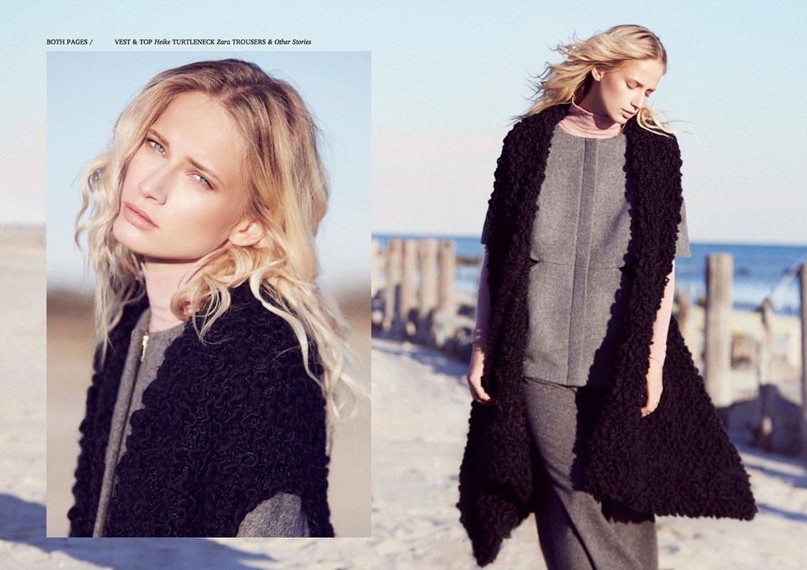 Fashionable blonde woman poses in grey clothing and black textured vest on a beach, featuring natural light and soft color grading.