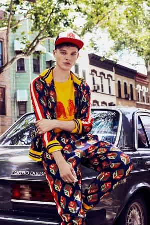 Young adult posing in patterned suit, cap, and yellow shirt, leaning on vintage Turbo Diesel car on urban street, sunny day.