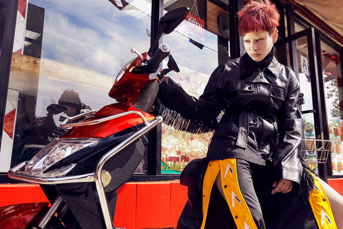 Stylish woman with red hair posing with a red and black scooter in front of a building with large windows, illuminated by natural light.