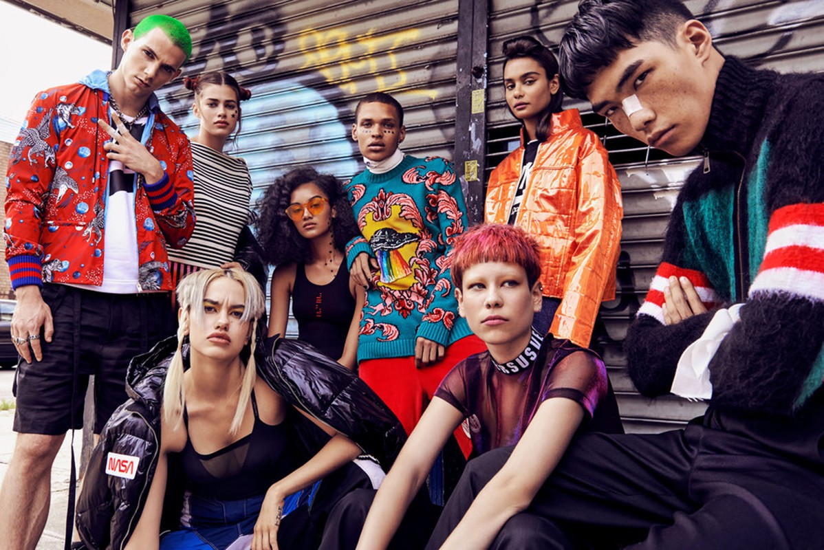Diverse group of young adults in vibrant streetwear poses outdoors against a graffiti-covered backdrop. Fashion-forward, colorful street style.