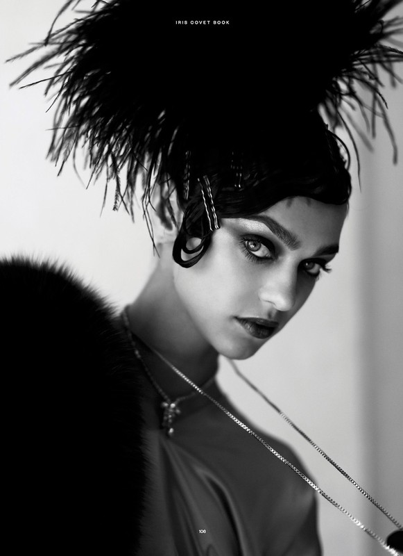 Monochromatic portrait of a woman with dark hair and dramatic eye makeup, wearing a feathered headpiece and a statement necklace.